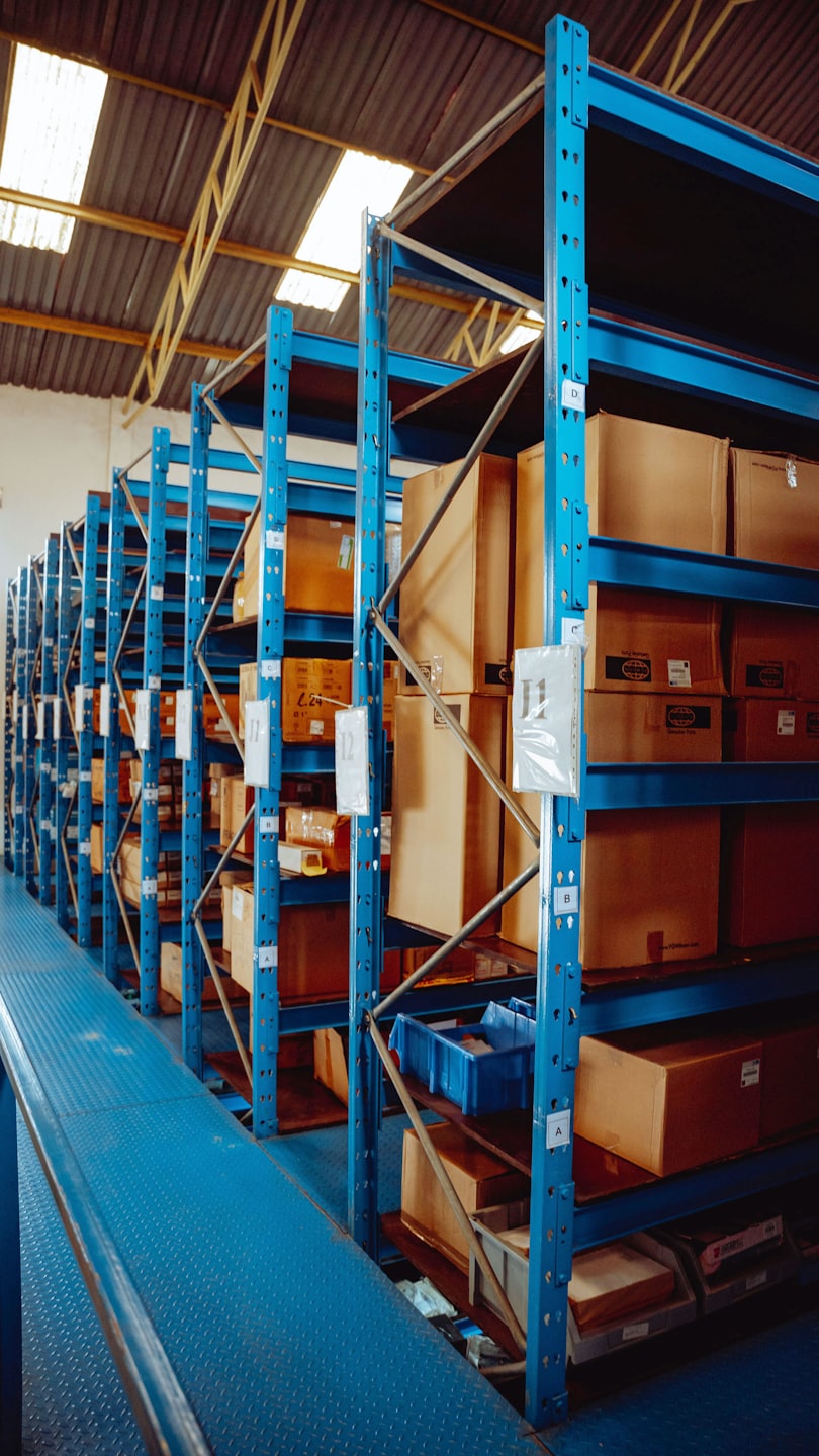 Pasillo de almacén con estanterías altas y azules apiladas con cajas.